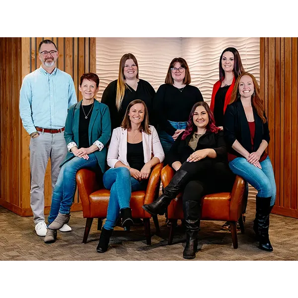 Six women posing for a professional team photo in a modern office lounge with orange chairs and wood paneling.