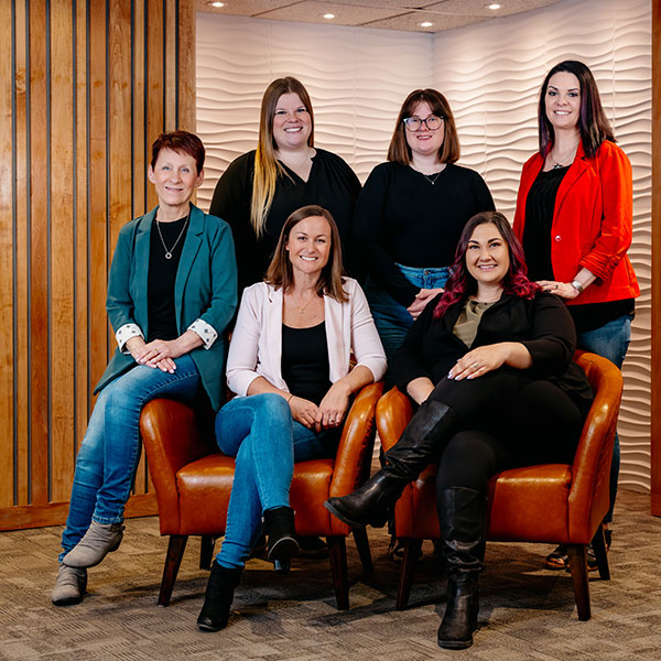 Six women posing for a professional team photo in a modern office lounge with orange chairs and wood paneling.