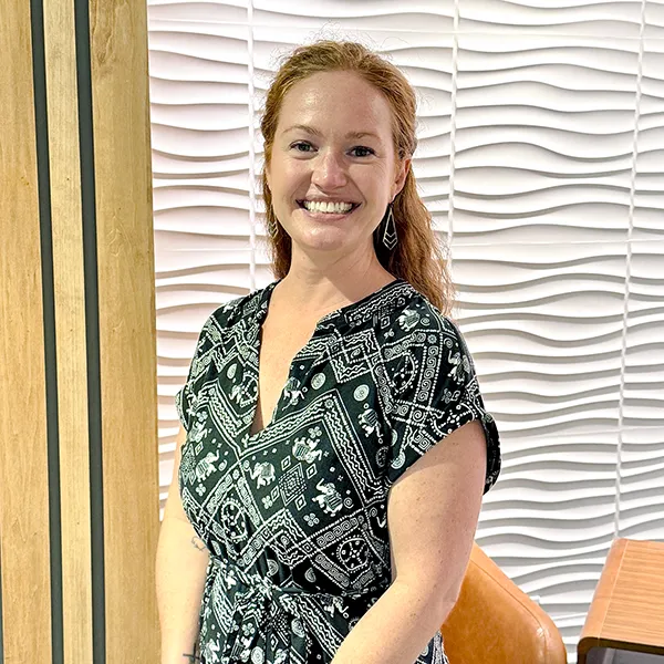 Smiling woman with red hair wearing a black-and-white patterned dress, standing indoors against a wavy white wall panel backdrop.